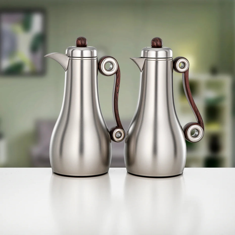 Two stainless steel coffee makers with dark brown wooden handles and matching dark brown lid knobs, displayed side-by-side on a white surface with a blurred green background.
